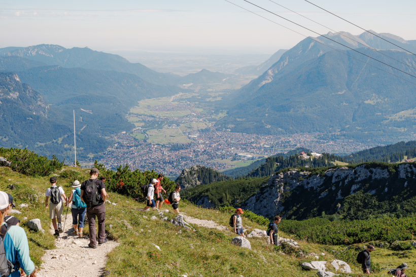 Group of hikers walking on a mountain trail overlooking a valley and distant town.