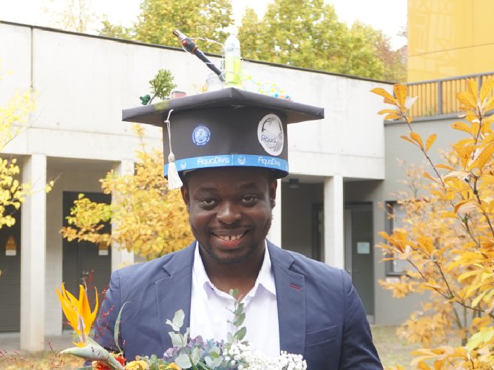 Graduate wearing decorated mortarboard cap with a bottle and other items, holding a bouquet outdoors.